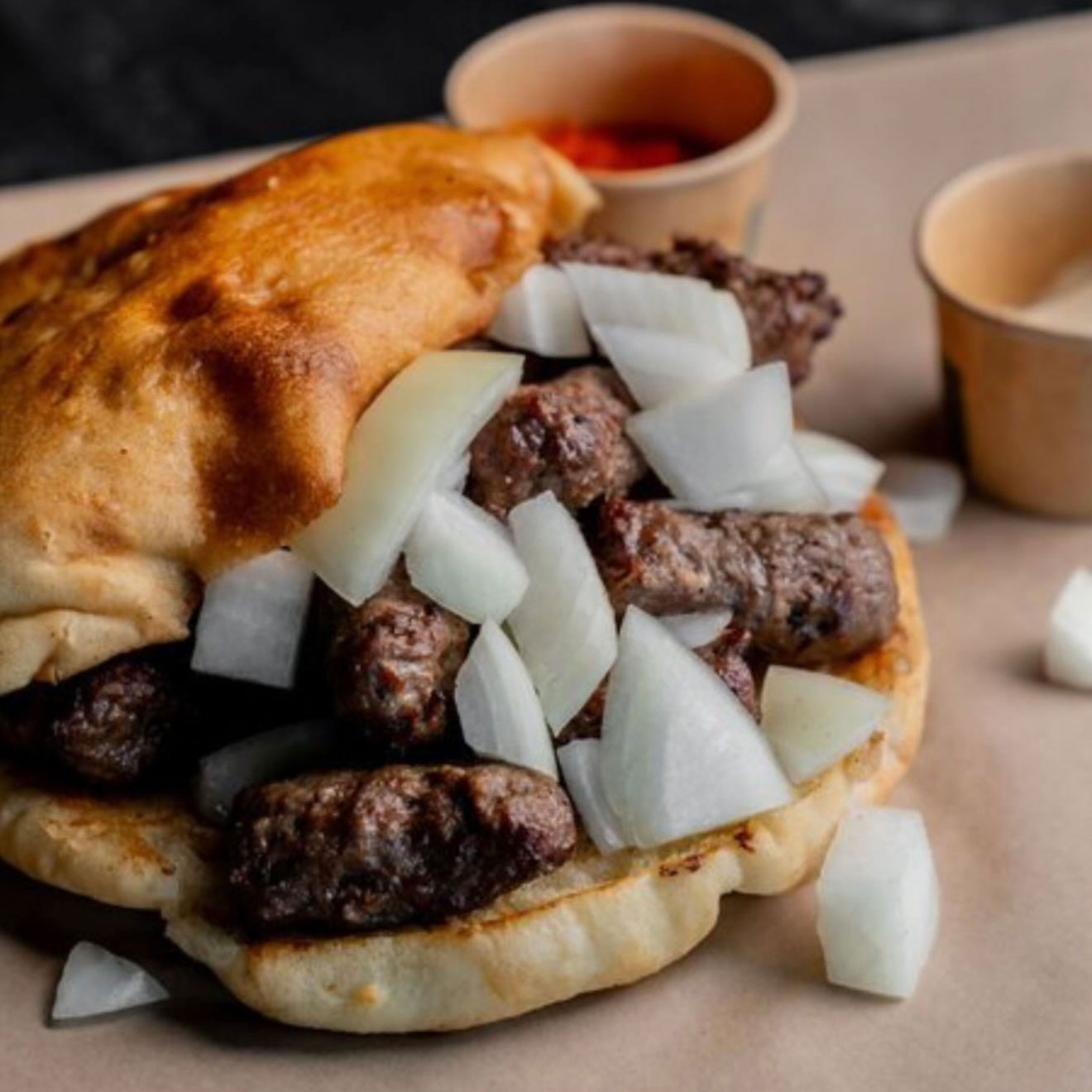 Ein Sandwich mit Ćevapčići, Zwiebeln und Fladenbrot, auf braunem Papier serviert.