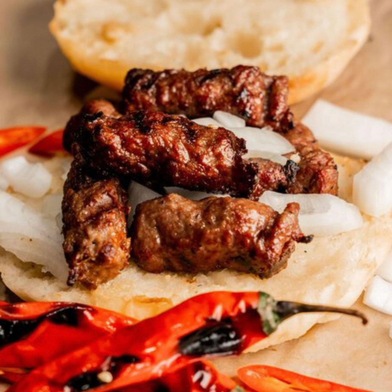 Nahaufnahme eines Cevapcici-Sandwiches mit roten und weißen Zwiebeln, im Vordergrund rote Chili.